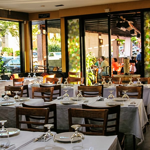 Bright restaurant dining room with set tables, large windows, and an outdoor patio with diners.