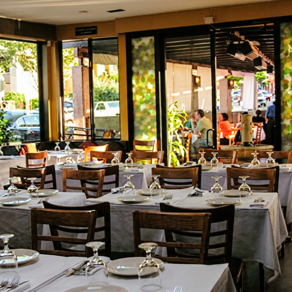 Bright restaurant dining room with set tables, large windows, and an outdoor patio with diners.