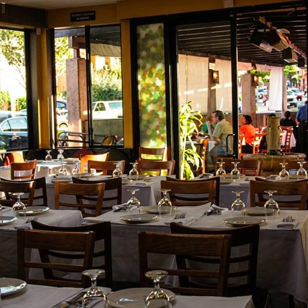 Sunny restaurant dining room with set tables, wooden chairs, and an outdoor patio with diners visible.