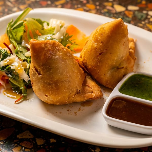Two golden samosas with a fresh salad and two dipping sauces on a white plate.